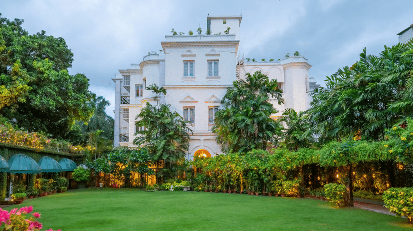 Facade view of Kenilworth Hotel, a city centre hotel in Kolkata, with a lawn in front of it, trees having fairy lights, many trees surrounding the lawn area as seen in late evening.