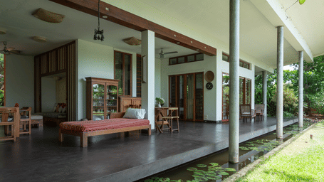 A backyard at Villa Wuwei in Mango Hill Aurovill, Pondicherry featuring a lush green garden, and an indoor area which features a bed, a seating area, a cupboard with utensils, etc.