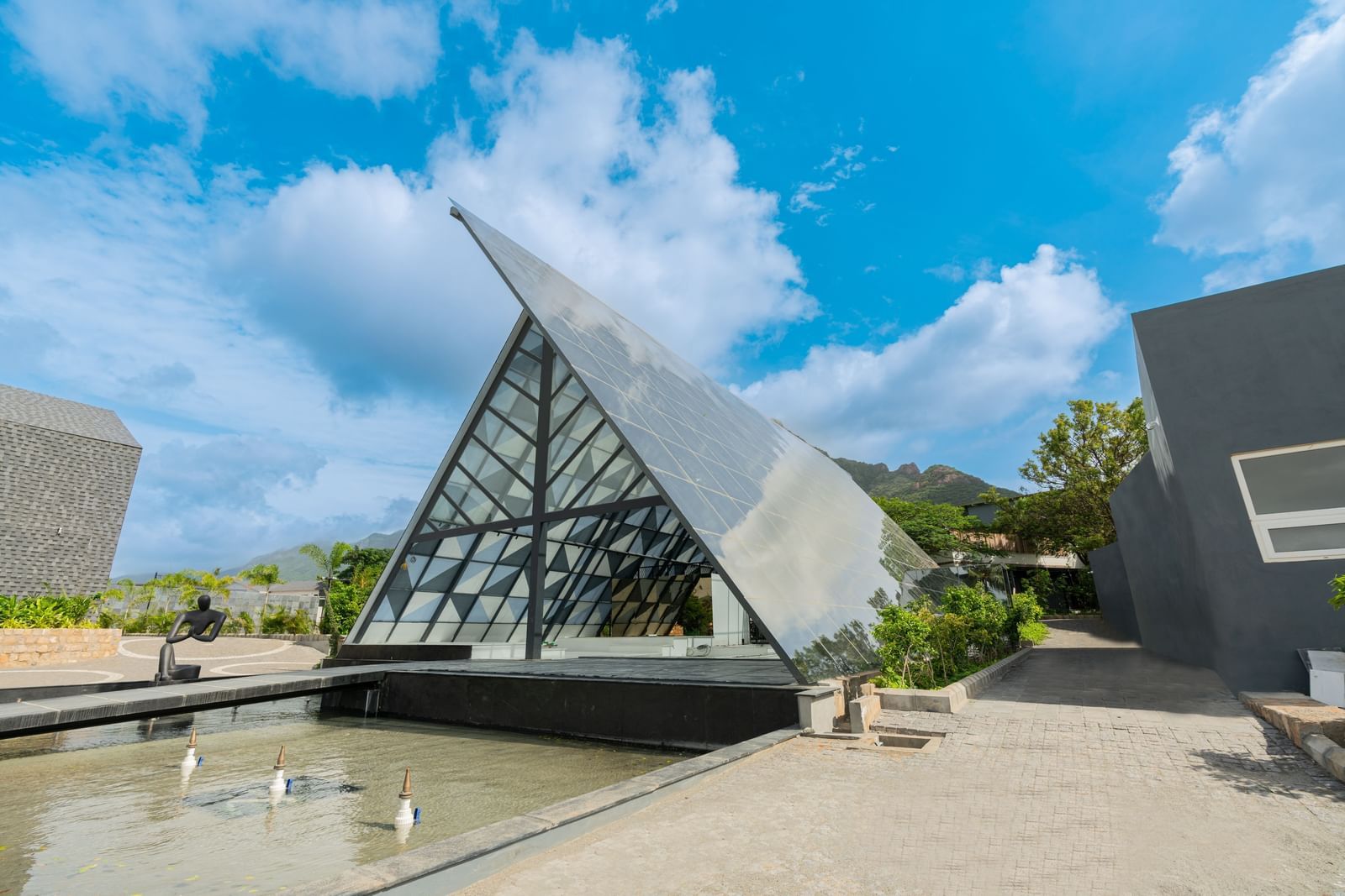 Exterior of the lobby of Darza Resorts in Coimbatore with a fountain pool in front of it and cloudy blue sky in the background.
