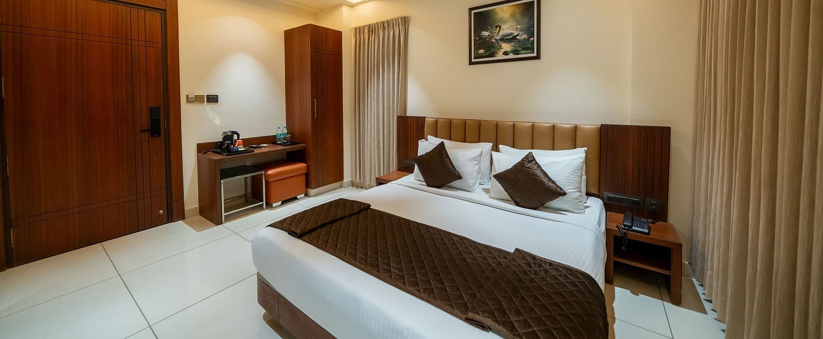 Hari Govinda's Express, Vijayanagar, features a neatly made bed with a photo frame above, a table and cupboard to the left, and drawn curtains to the right.