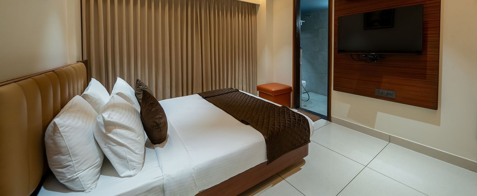 A side view of the bed at Hari Govinda's Express, Vijayanagar, with a photo frame above the headboard, fully drawn curtains, a doorway, and a wall-mounted TV beside it.