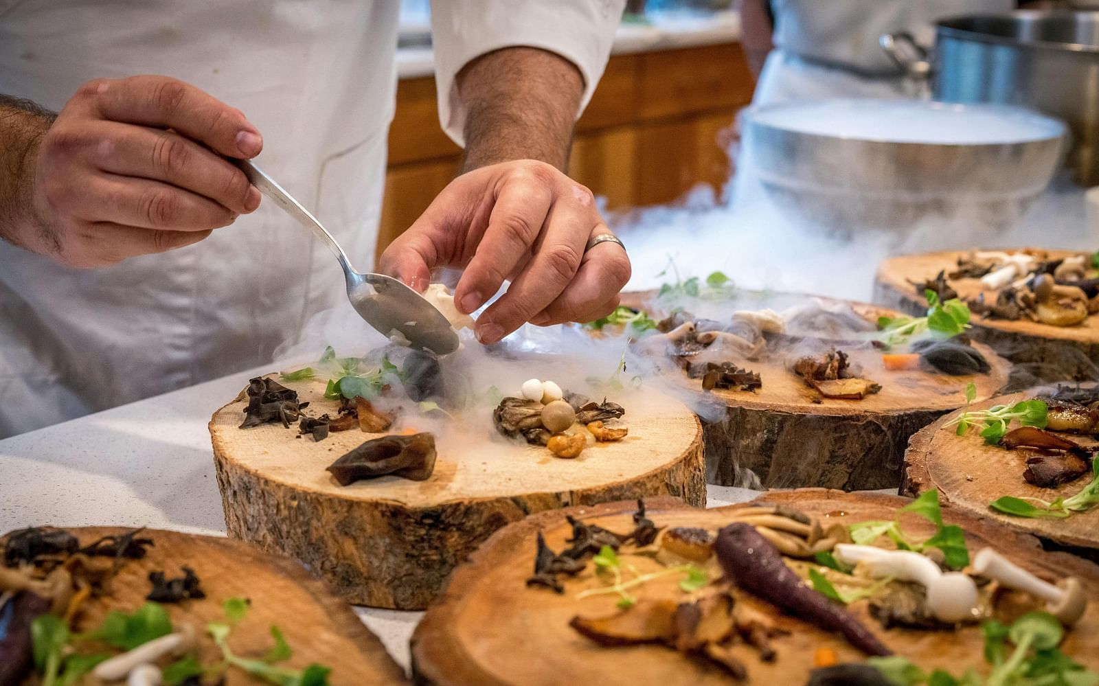 A chef's hands are meticulously plating small, intricate dishes on wooden serving platters, surrounded by various ingredients and garnishes.