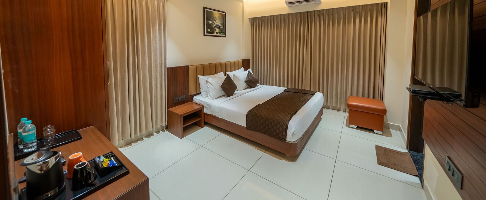 A spacious room at Hari Govinda's Express, Vijayanagar, featuring a large bed with a wooden headboard, a bedside table, a wall-mounted TV, and a wooden table with essentials, with curtains fully drawn to the side.