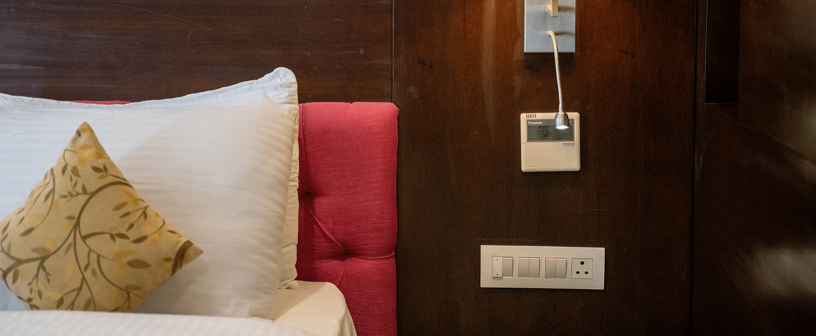 A cosy hotel bed with white linens, decorative pillows, and a bedside lamp on a wooden panel - Hotel Udai Median, Udaipur.