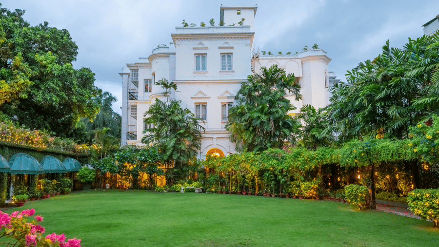 Facade view of Kenilworth Hotel, a city centre hotel in Kolkata, with a lawn in front of it, trees having fairy lights, many trees surrounding the lawn area as seen in late evening.
