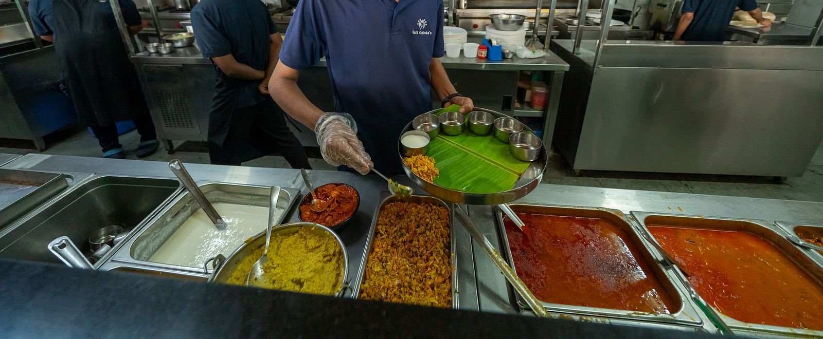 A person serving food from the counter area with one hand while holding a plate in the other at Hari Govinda's