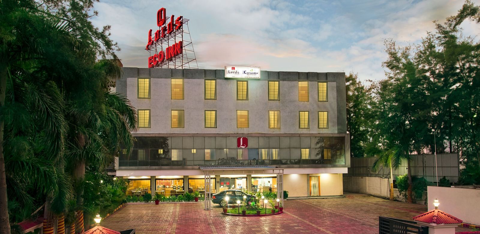The exterior of a modern hotel with a red and white flag and landscaped entrance - Lords Eco Inn Dahej