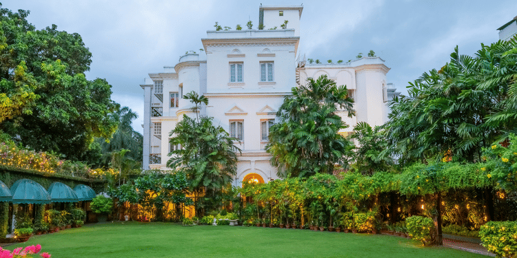 Facade view of Kenilworth Hotel, a city centre hotel in Kolkata, with a lawn in front of it, trees having fairy lights, many trees surrounding the lawn area as seen during twilight hour.