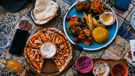 Overhead view of a vibrant meal with a sliced pizza, grilled meat platters, rice, and colourful side dips.