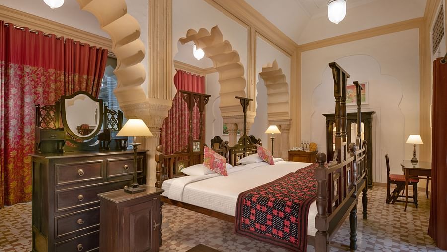 A room with a four-poster bed, a dresser with a mirror, a chair, and arched doorways with decorative columns in Tijara Fort-Palace - 19th Century, Alwar.