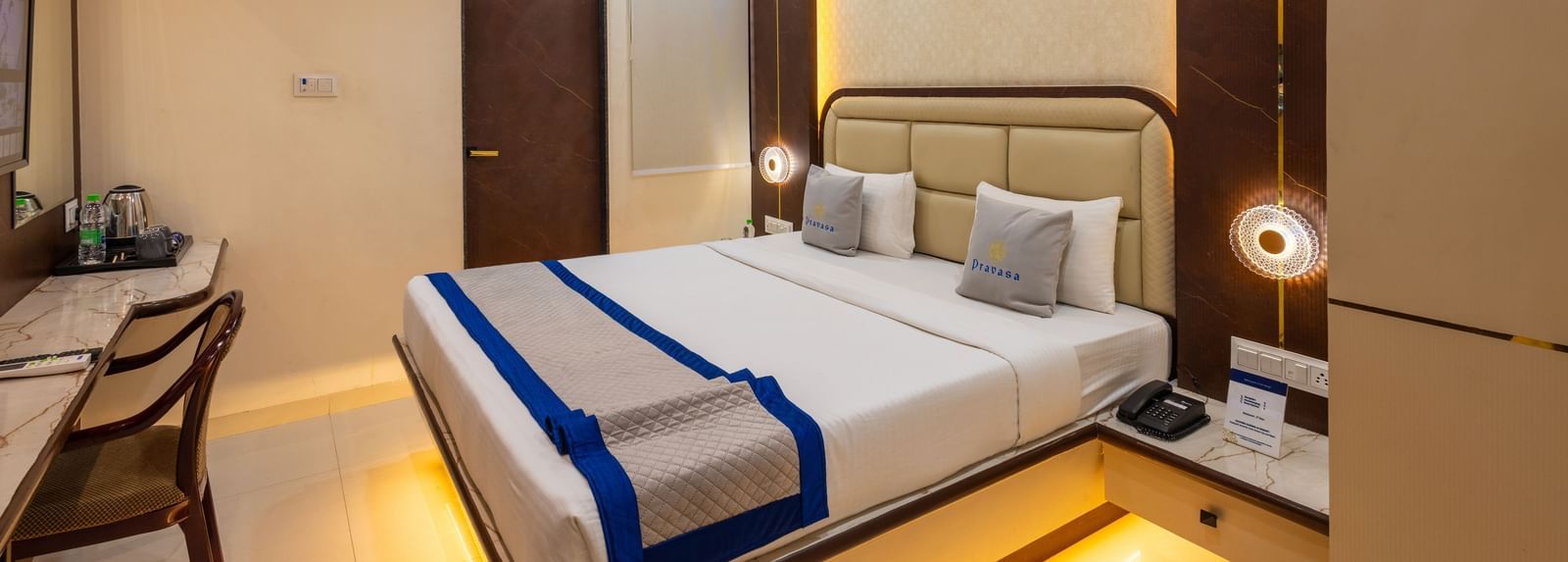 A room with a double bed, warm lighting, blue & beige runner, bedside lamps & a chair facing the wall at Pravasa Stay Dewas Naka, Indore.