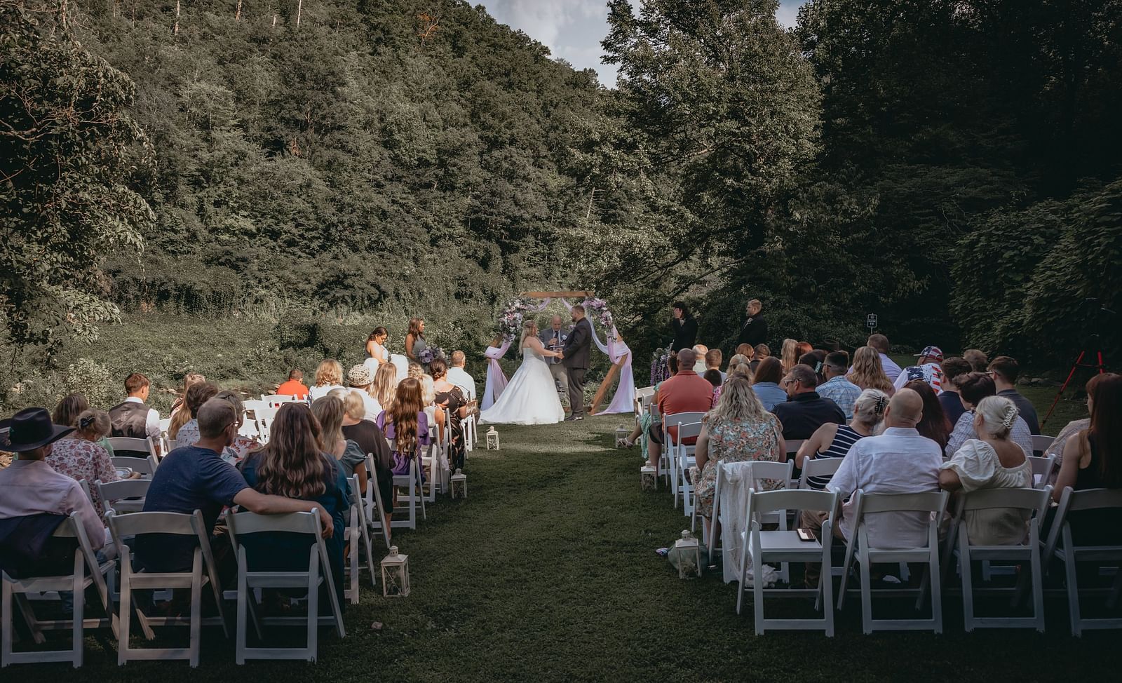 Couple taking their wedding vows near the wooden decorated frame at the Historic Tapoco Lodge. On either side of the aisle people are seated to attend the wedding.