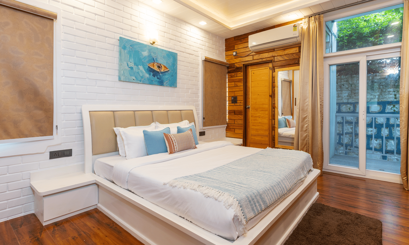 A modern bedroom with a large bed, light wood panelling, a glass door to the outside, and a bright blue artwork - Megha Resort & Villas, Dehradun