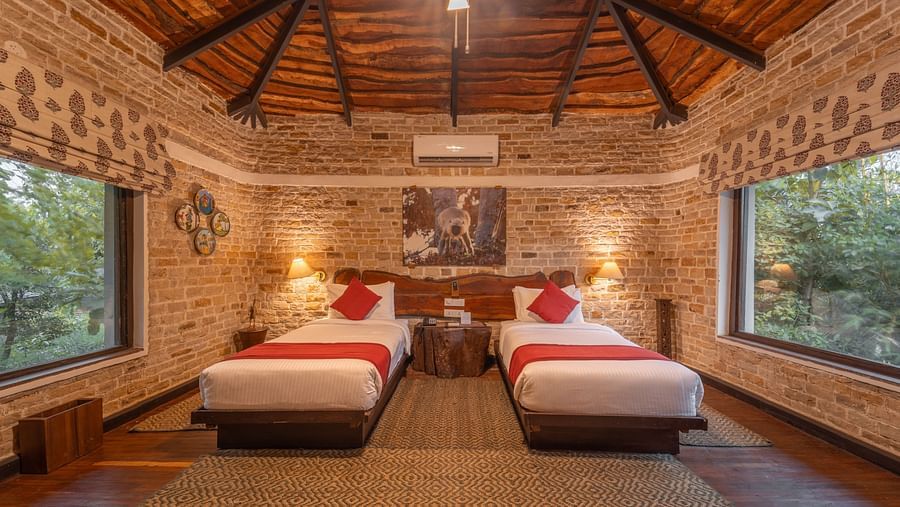 A bedroom at Tendu Leaf Jungle Resort featuring twin beds, large window on wither side , a bedside table, and a ceiling light in a wooden furnished room.
