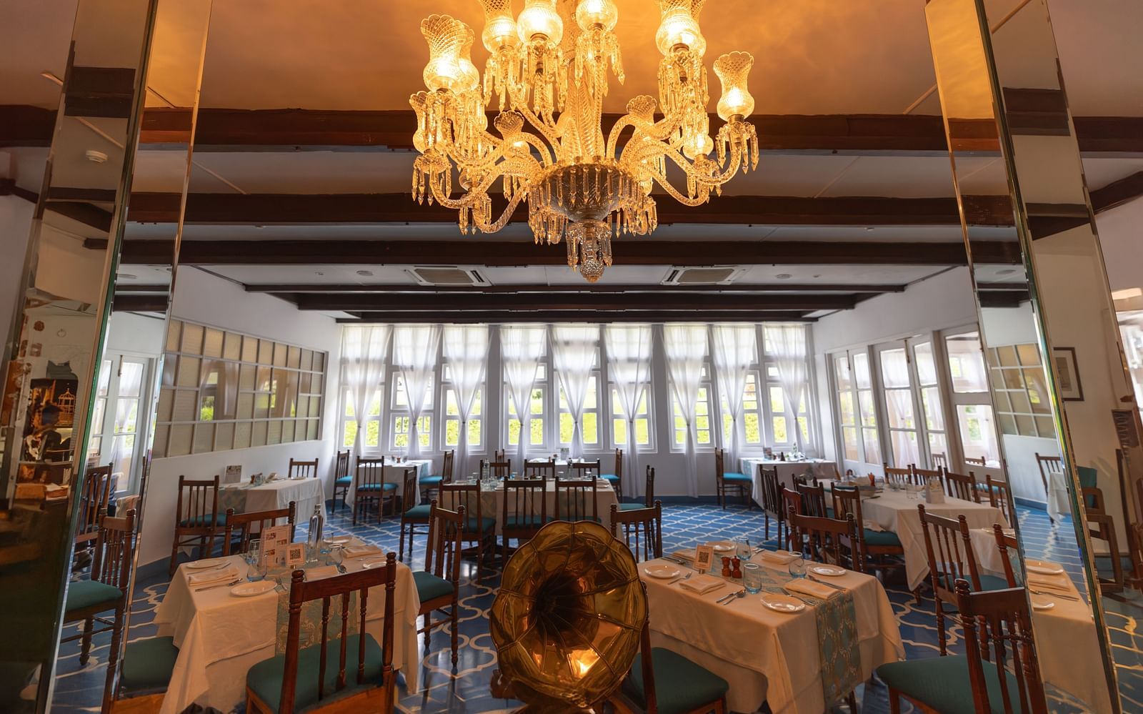 Dining arrangement in a restaurant at Saraca Hotel, Lucknow with large French windows in the backdrop and a chandelier hanging from the centre of the ceiling