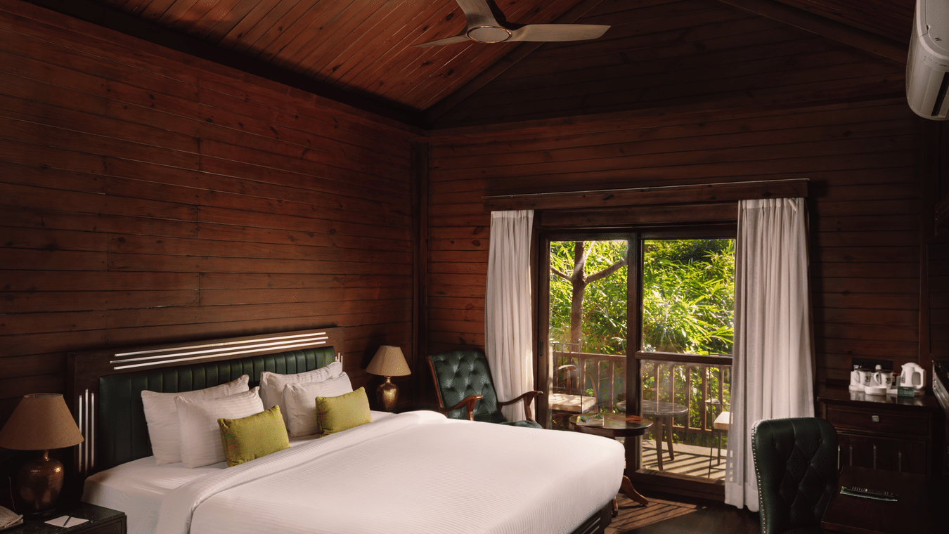 A bedroom at The Ananta Udaipur featuring a king size bed, 2 side tables, armchair, desk, and sliding glass doors leading to a balcony.