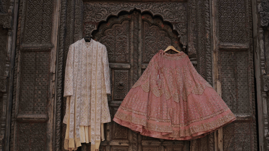 Elegant bridal lehenga and groom sherwani displayed against an intricately carved heritage doorway ahead of a wedding celebration.