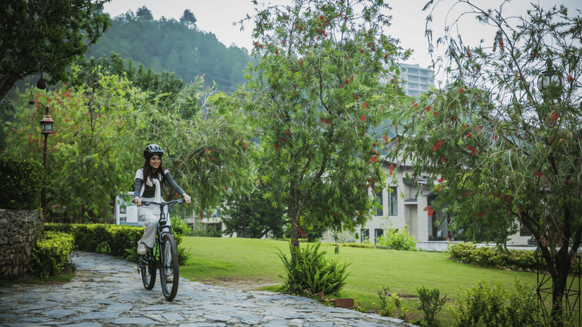 A woman riding a bicycle on stone pathway bordered by lush garden plants, with misty hills in the background, showcasing wellness and recreational activities in the scenic environment of The Manor Sports & Wellness Hotel in Shimla.