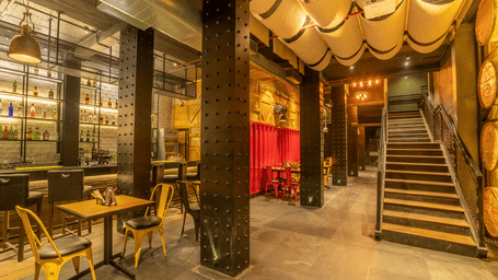 The interior of a dimly lit bar shows tables, chairs, and a staircase leading to an upper level at S Hotels, Chennai.