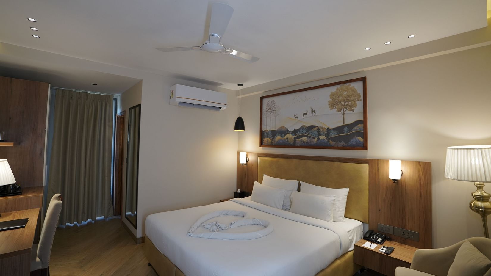 A warm-lit room featuring a king-size bed with a painting above the headboard and an AC mounted next to it