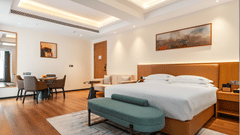 Spacious bedroom at Bamus Sarovar Portico, Kampala, with a large bed, a bench, a dining area, and recessed ceiling lighting.