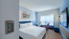 A bright hotel room contains a neatly made bed with white linens, a bedside table, and a window with curtains | Standard Room | Sarovar Portico, Sriperumbudur