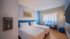 Bright, modern hotel room with two twin beds, a small seating area by a window with sheer curtains, and a wall-mounted water dispenser.