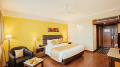A modern bedroom with a neatly-made bed, yellow accent wall, leather armchair, coffee table with a fruit plate, and wooden flooring at The Piccadilly Lucknow.