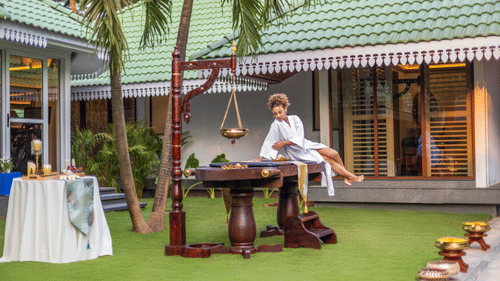 A woman in bathrobe getting on a spa bed which is placed in a lawn of Heritage Village Resort and Spa, Goa. The lawn also has other spa products. 