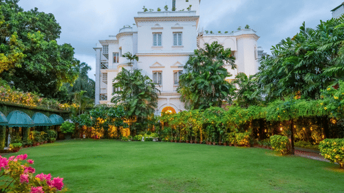 Facade view of Kenilworth Hotel, a city centre hotel in Kolkata, with a lawn in front of it, trees having fairy lights, many trees surrounding the lawn area as seen during twilight hour.