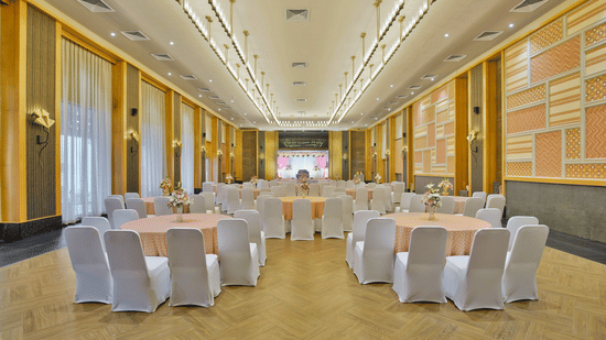 A wide, brightly lit fortress hall prepared for a formal event. The hall features high ceilings with elegant paneling, and several round tables set with linens and matching chair covers Pramod Hotels & Resorts. 