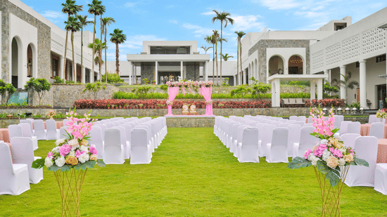 An outdoor wedding venue setup on a lush green lawn, featuring rows of chairs facing a floral archway, with a large, modern white building and palm trees in the background under the sky at Pramod Hotels & Resorts.