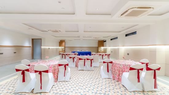 Banquet Hall at Pride Comfort Dwarka featuring chairs and table decorated with ribbons and table cloth respectively. There is also a conference table with chairs at one end.