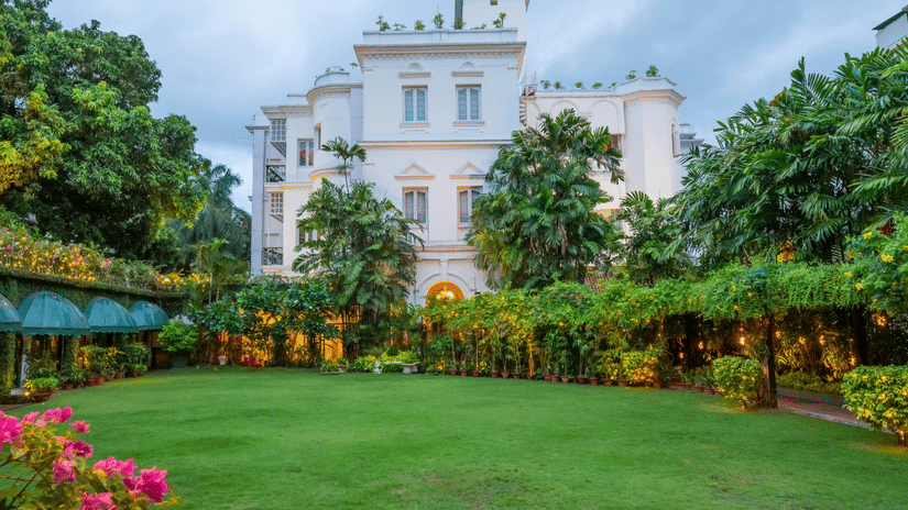 Facade view of Kenilworth Hotel, a city centre hotel in Kolkata, with a lawn in front of it, trees having fairy lights, many trees surrounding the lawn area as seen during twilight hour.