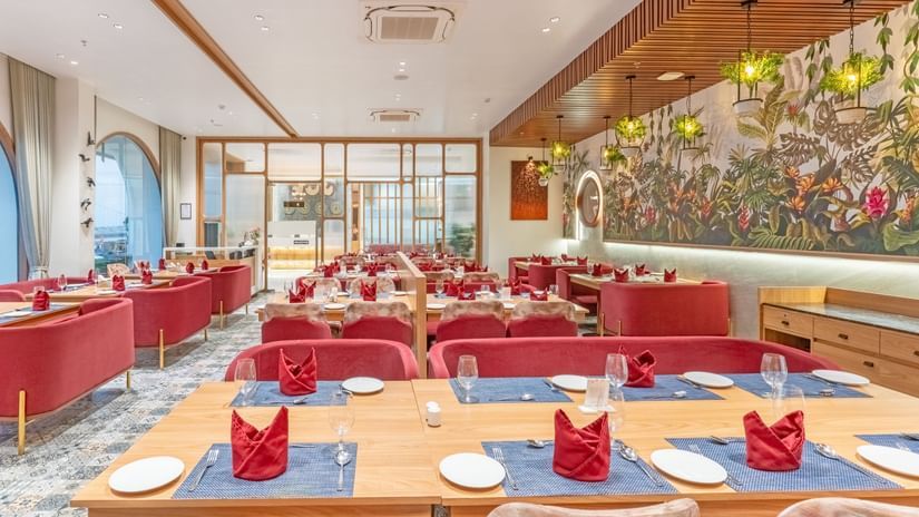 A restaurant at Pride Comfort Dwarka featuring chairs and tables, glass walls, and flower pots with lights hanging from the ceiling.