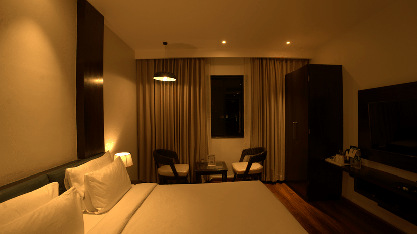 A room at Wedlock Greens Hotels & Resorts, Dhanbad featuring a double size bed, a flat TV screen mounted on the wall, a seating area with chairs and centre table and a small window.