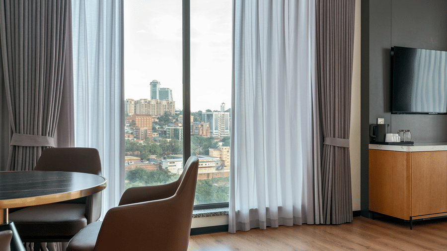 Interior view at Bamus Sarovar Portico, Kampala, showing a dining table by a large window with city views and curtains.