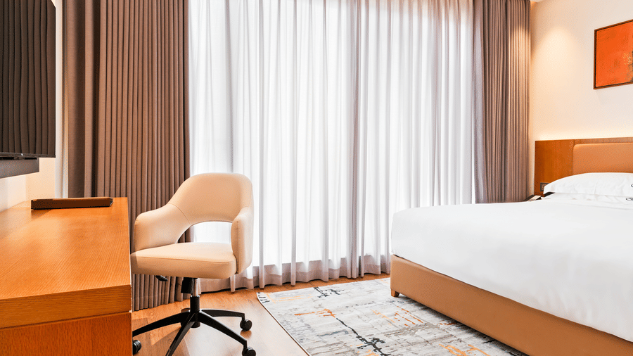 A wooden desk with a chair positioned next to a bed with linens and a patterned rug at Bamus Sarovar Portico, Kampala.