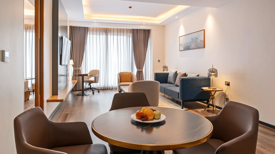 A round dining table with 4 brown chairs leading into a living area with a sofa at Bamus Sarovar Portico, Kampala.