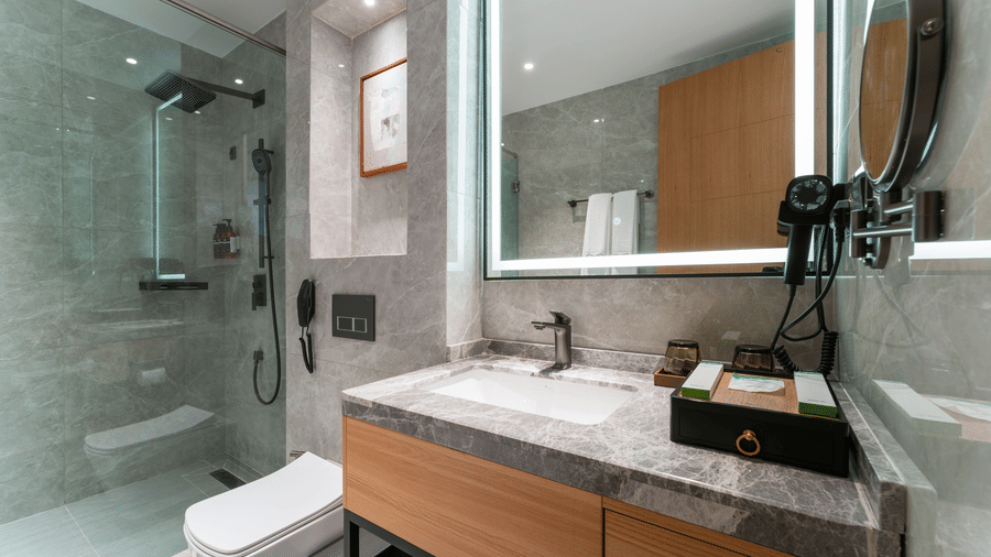 A modern bathroom with a marble vanity, a backlit mirror, a walk-in shower, and a toilet at Bamus Sarovar Portico, Kampala.
