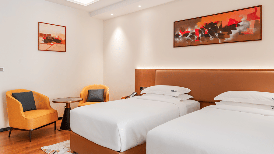 Twin bedroom at Bamus Sarovar Portico, Kampala, showing twin beds, upholstered armchairs, a small table, and framed artwork on the walls.
