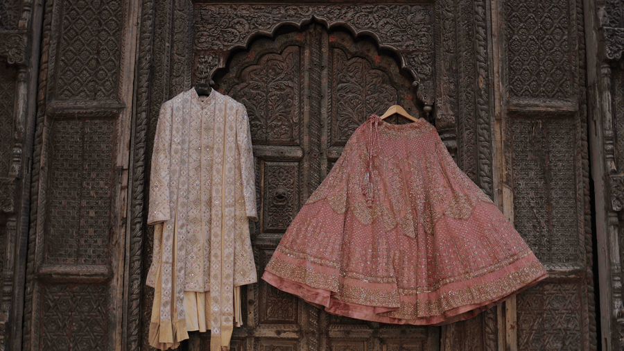 Elegant bridal lehenga and groom sherwani displayed against an intricately carved heritage doorway ahead of a wedding celebration.