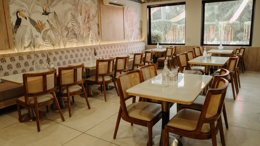 Indoor dining area at Corbett Adventure Resort, Ramnagar featuring upholstered bench seating, cane chairs, wall art, and large windows.