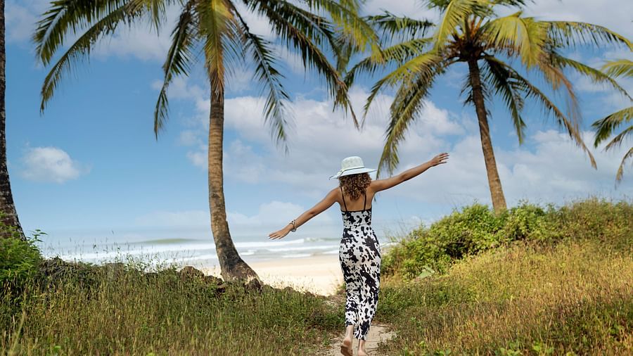 Woman walking through tall grass under palm trees towards the beach on a sunny day.