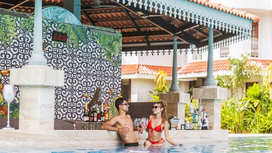 A man and a woman in the pool with a deck in the background with bottles lined up and resort in the background. 