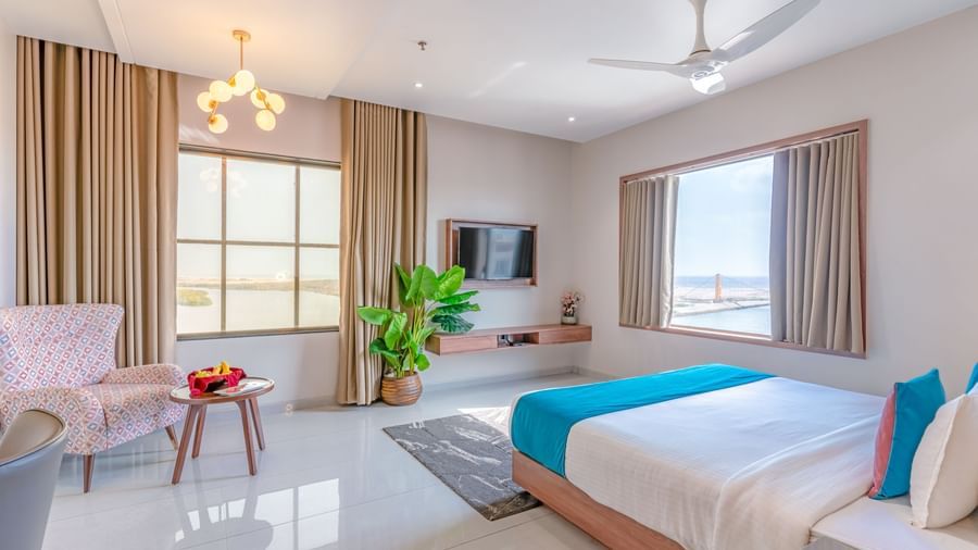 Suite at Pride Comfort Dwarka featuring a bed, two windows, flat TV screen, a sofa chair with centre table, a flat TV screen, lights hanging from the ceiling, and a flower pot.