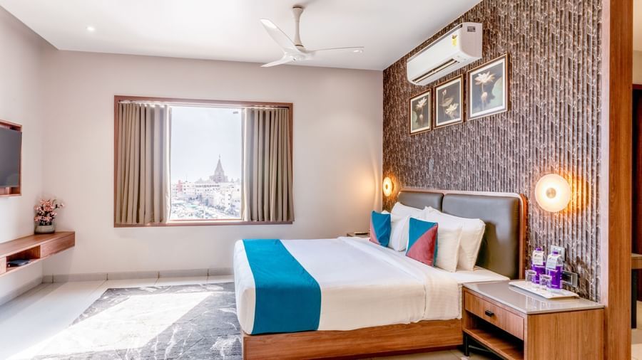 Suite Room at  Pride Comfort Dwarka featuring a bed, a bedside table with lamp, photo frames on the wall, an AC, a flat TV screen, and an open window.