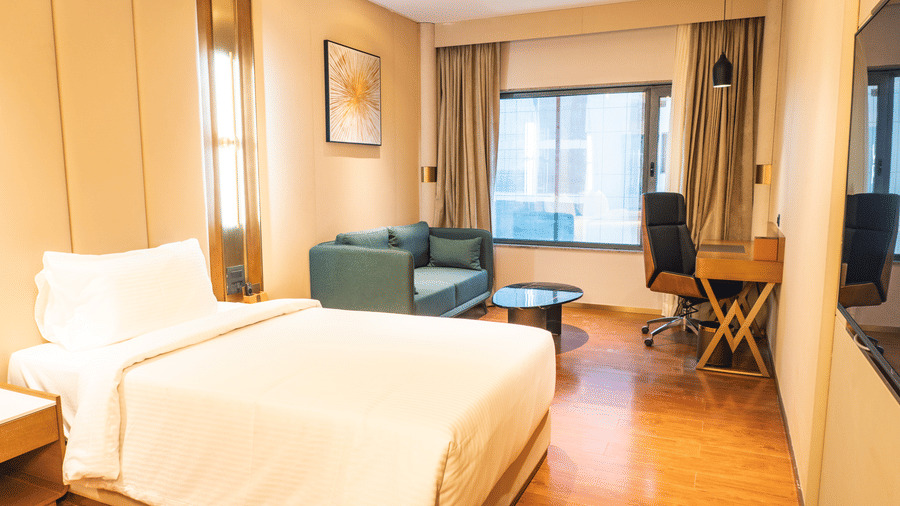 A king size bed, green sofa chair, and wooden study desk with a wheeled chair under a ceiling light at Pride Plaza Indore.