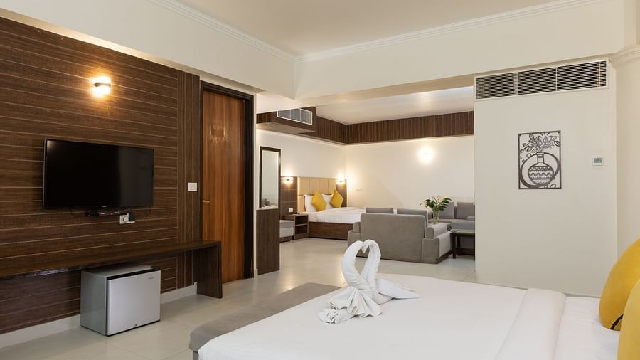 A spacious hotel bedroom with a large bed, featuring two towels folded into the shape of swans, a television, and a glimpse of another room beyond - Pride Premier Dehradun