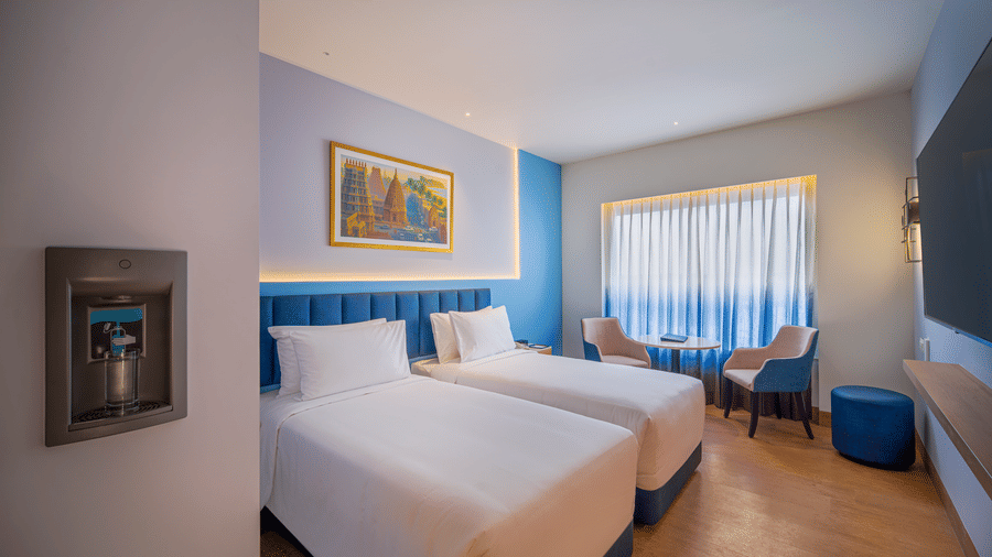 Bright, modern hotel room with two twin beds, a small seating area by a window with sheer curtains, and a wall-mounted water dispenser.